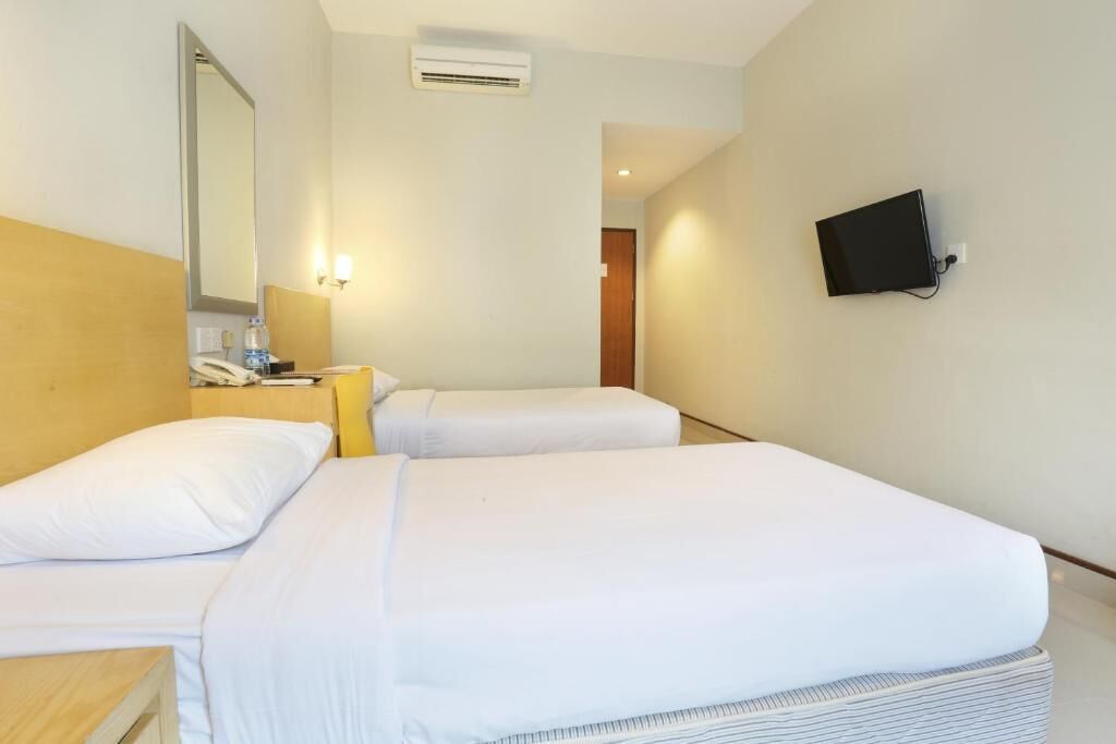 A hotel room with two beds, a bedside table with a lamp, a mirror, an air conditioning unit, and a TV mounted on the wall.