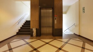 A modern elevator in a hallway with a checkered floor pattern, a trash bin, and a stairwell with a handrail. - Triniti Hotel Batam (Batam)