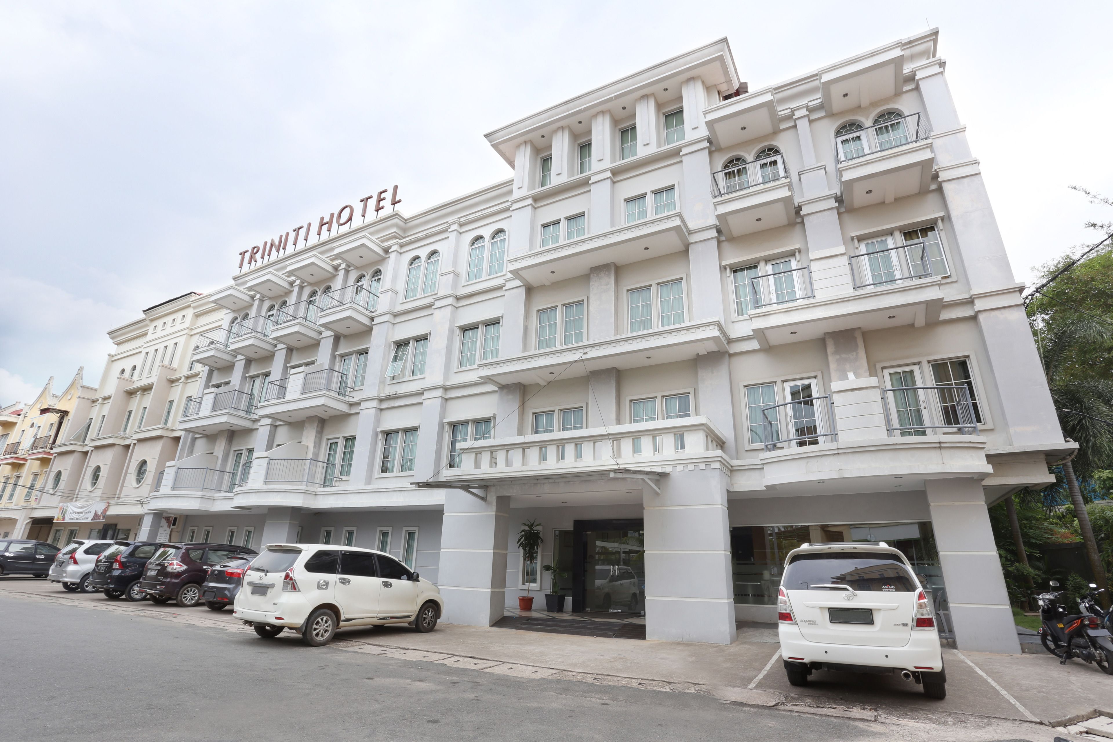 A multi-story hotel with a white facade, balconies, and a sign reading "TRINITI HOTEL".