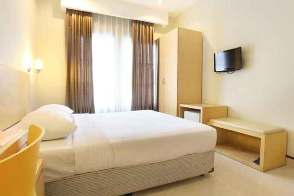 A hotel room with a large bed, a bedside table, a TV mounted on the wall, and a window with curtains.