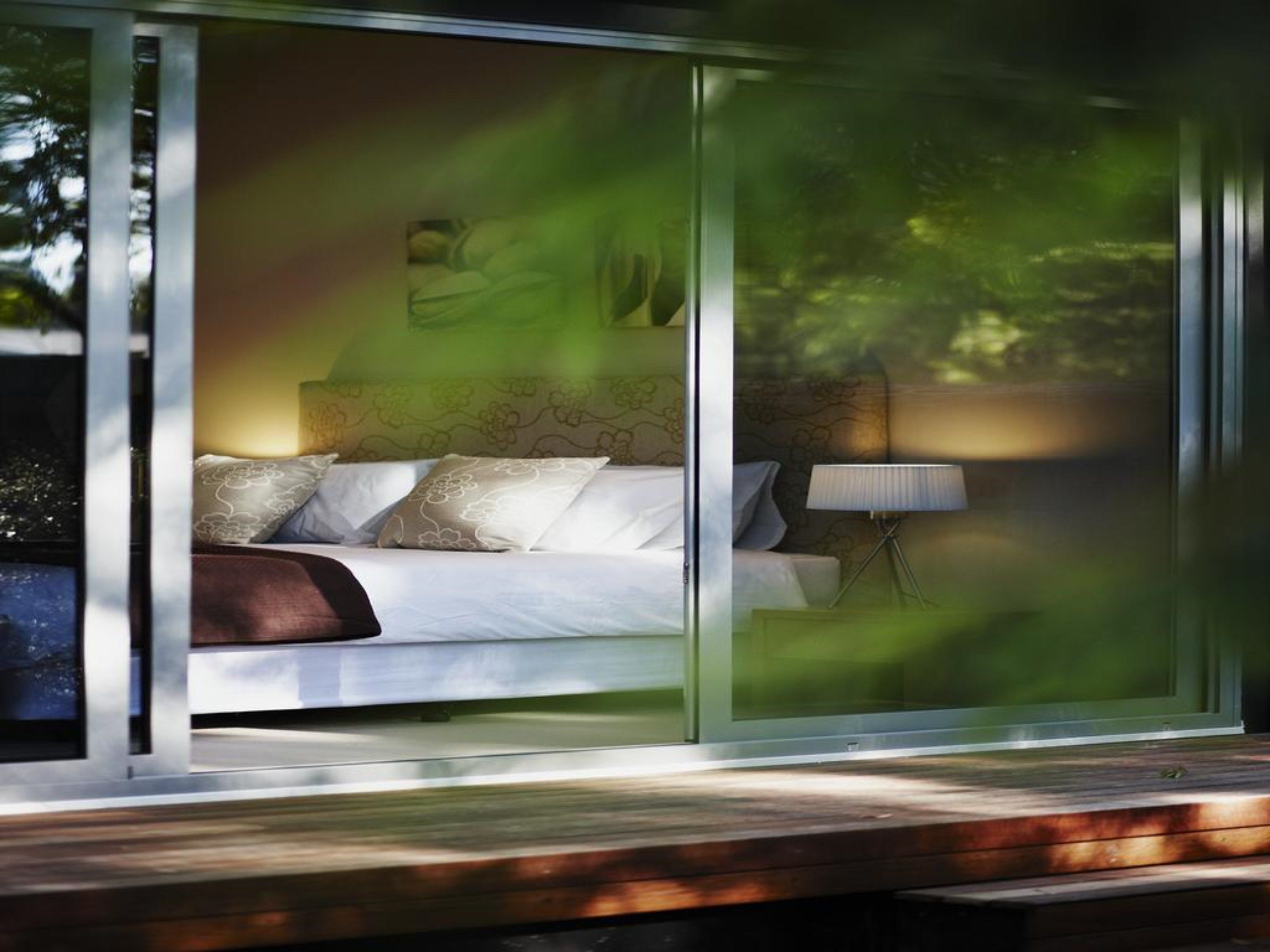 A modern bedroom with a large bed, a bedside lamp, and a view of greenery outside.