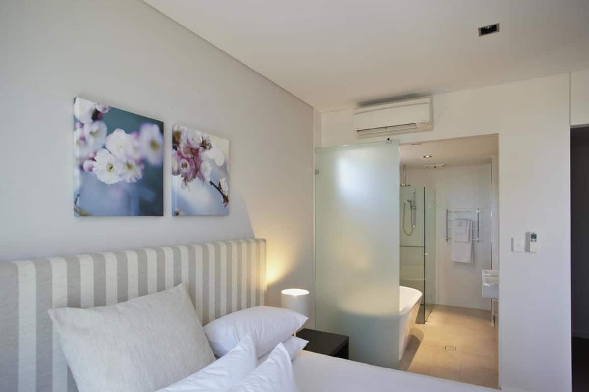 A hotel room with a bed, a nightstand, a wall-mounted air conditioner, and a bathroom visible through a glass door.