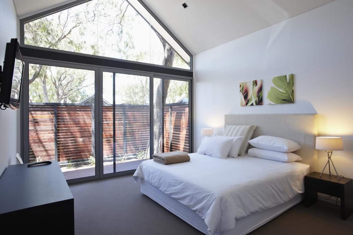 A modern bedroom with a large window, a bed with white linens, a bedside table with a lamp, and artwork on the wall.