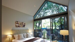 A modern bedroom with a large bed, bedside lamps, a desk with a lamp, and a view of trees through large windows. - Aqua Resort Busselton (Broadwater)