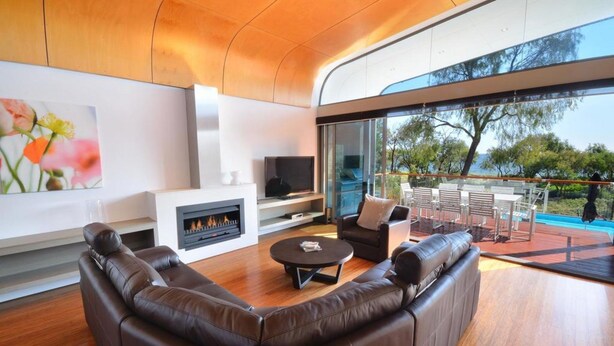 A modern living room with a fireplace, leather sofa, and a large glass door leading to an outdoor patio. - Aqua Resort Busselton (Broadwater)