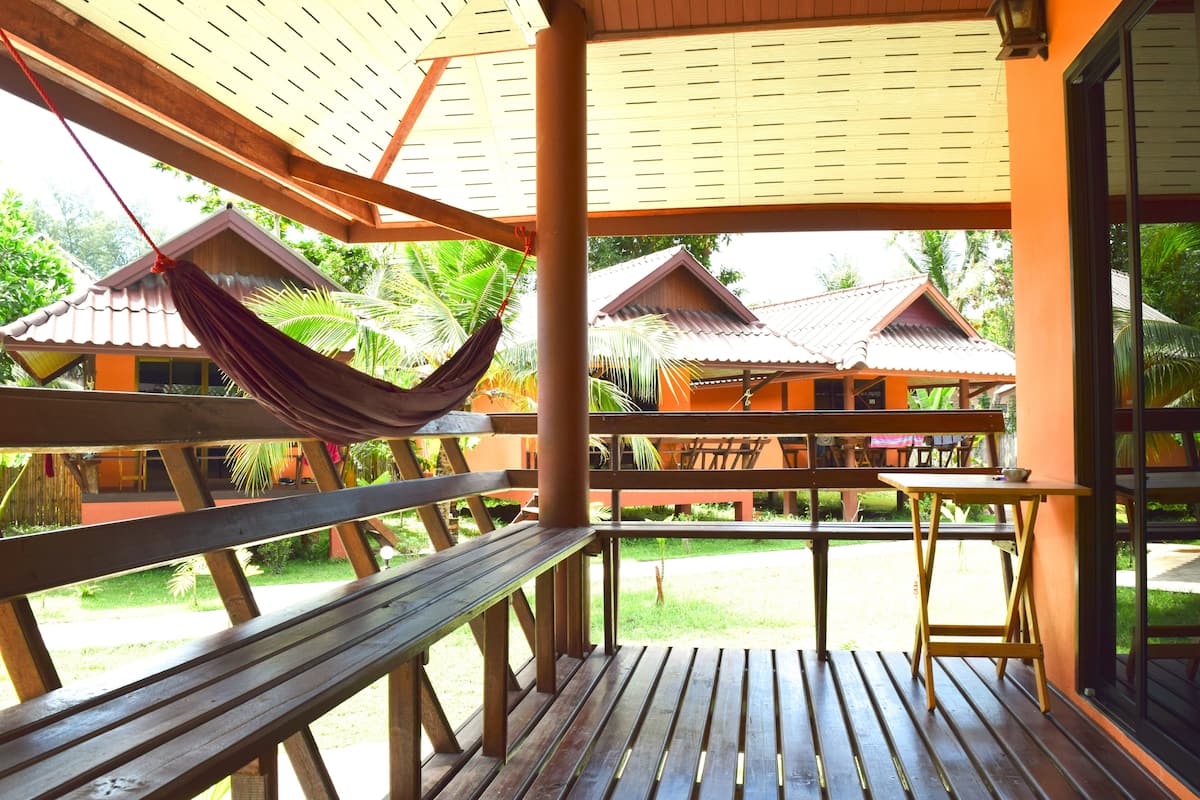 Une terrasse en bois, dotée de bancs et d’un hamac, qui offre une vue sur une cour intérieure agrémentée de bâtiments de style traditionnel et de palmiers.