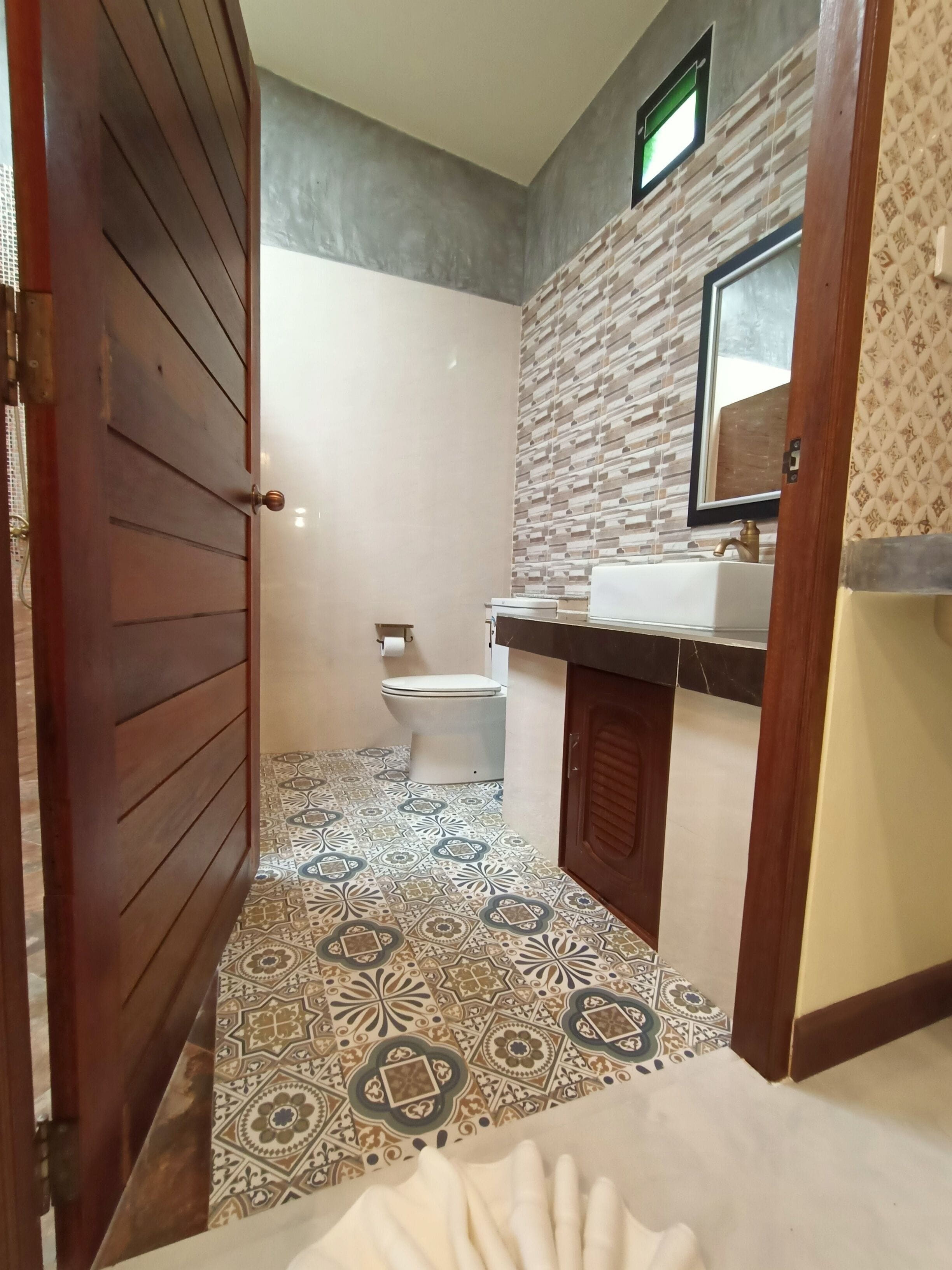 A bathroom with a patterned floor, a toilet, a sink with a mirror, and a wooden door.