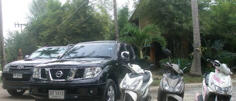A black SUV parked on a dirt road with a white scooter and a pink scooter beside it.