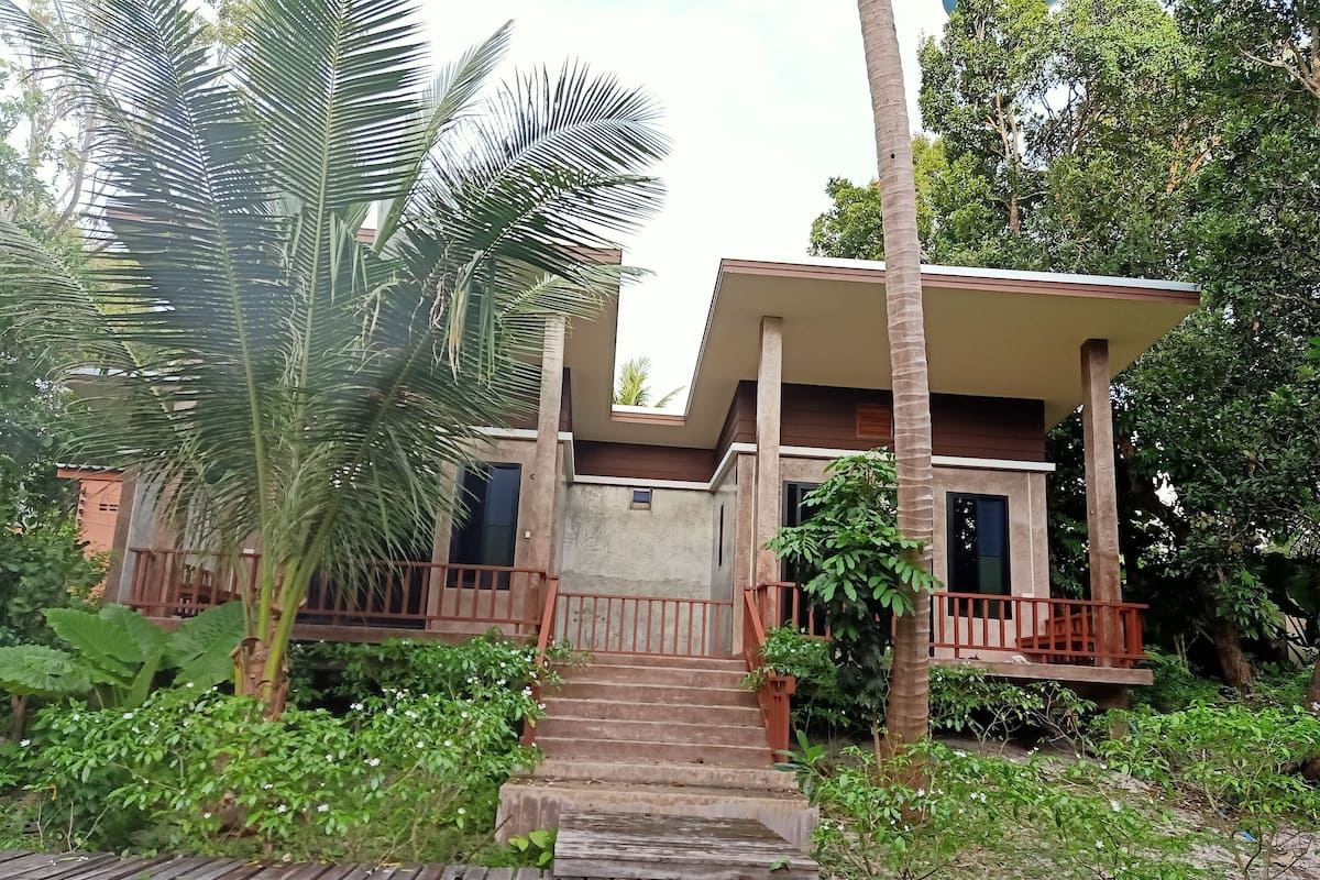 Une maison moderne dotée d’une terrasse en bois, entourée d’une végétation luxuriante et de palmiers.