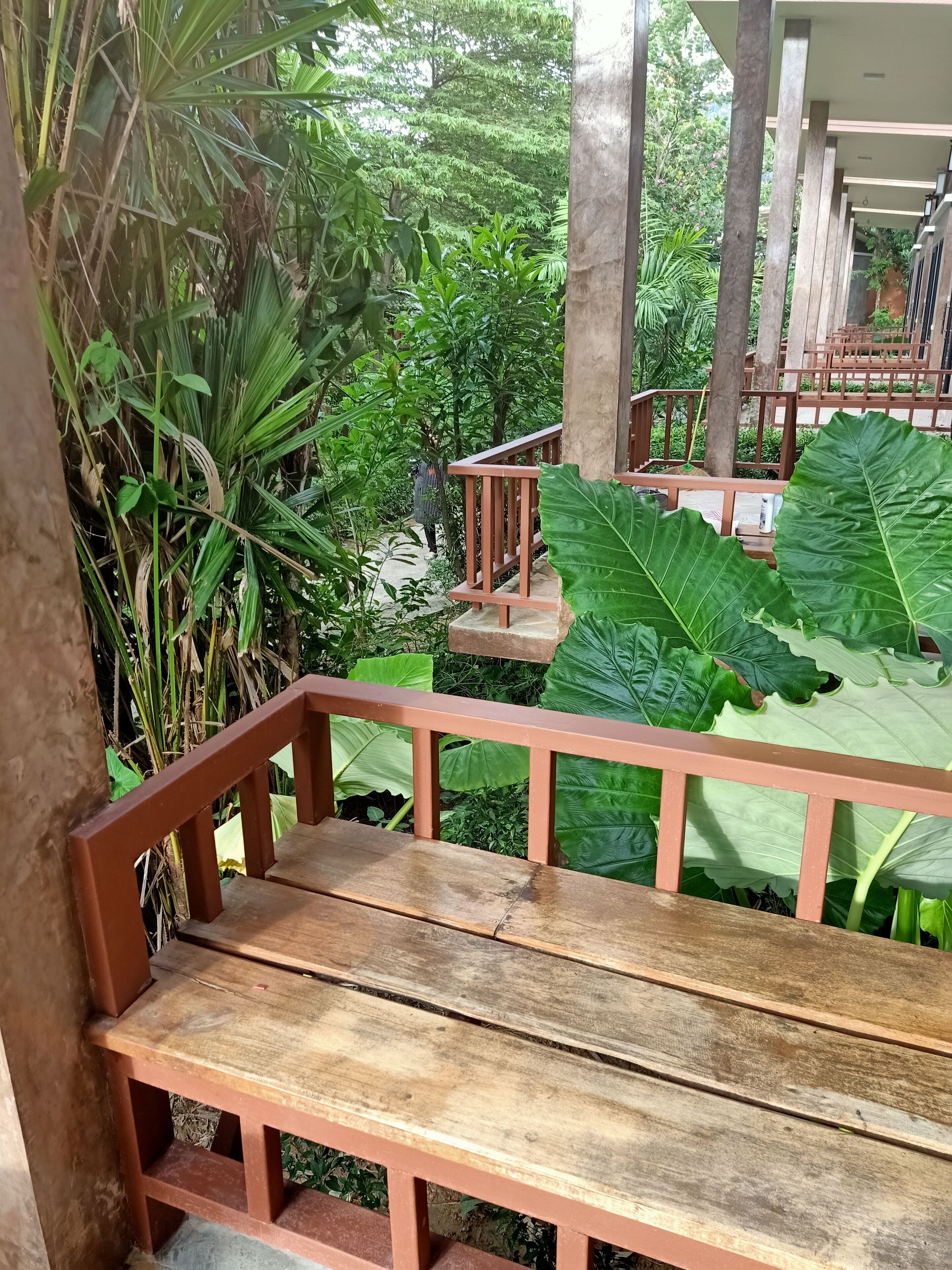 A wooden bench with a metal railing, surrounded by lush green foliage and a view of a building in the background.