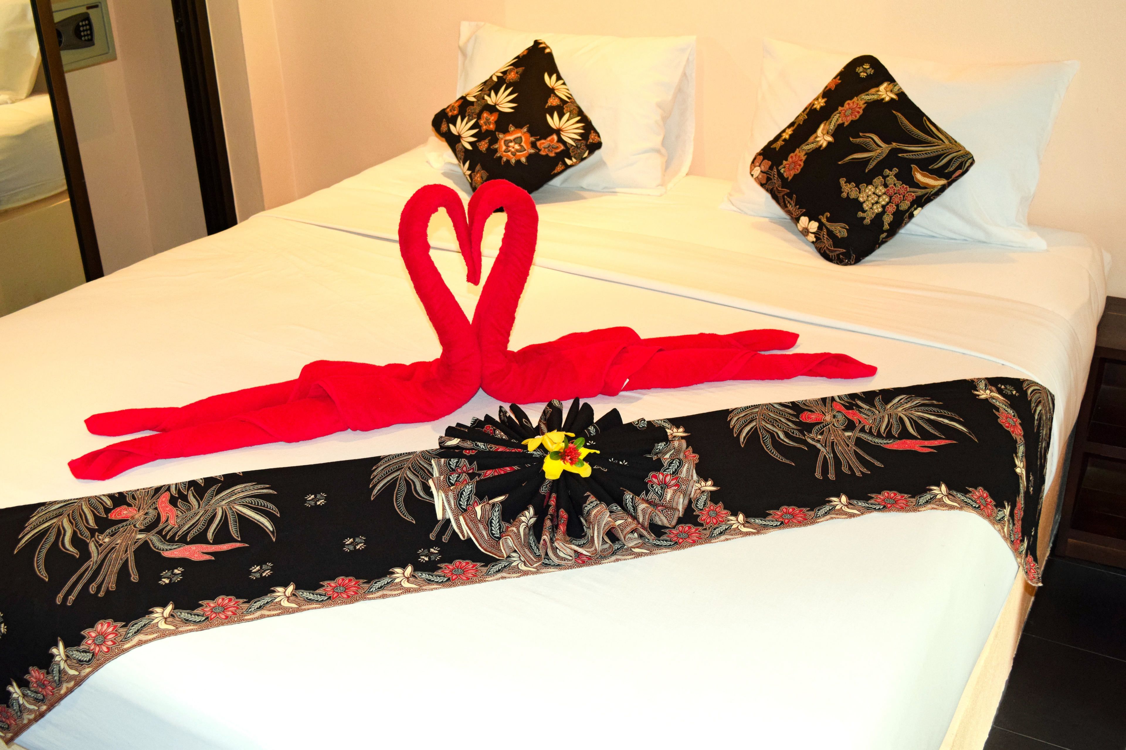 A neatly made bed with a red towel arranged in a heart shape, a black and red floral bedspread, and two decorative pillows.