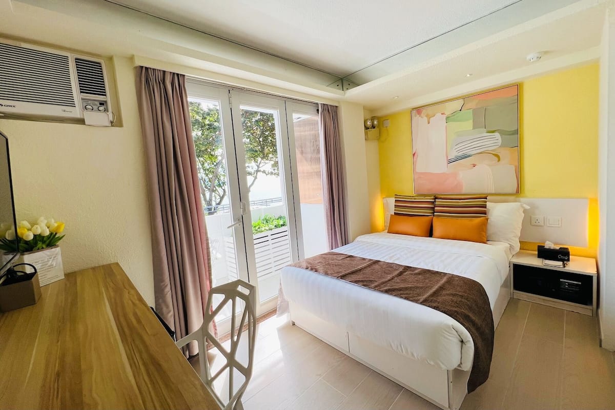 A hotel room with a bed, a desk, a chair, a window with curtains, and a wall-mounted air conditioner.