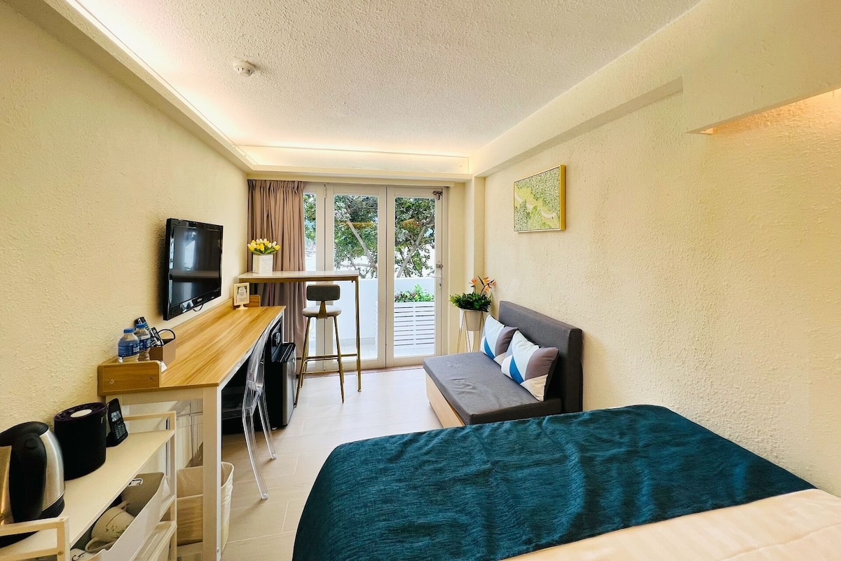 A compact hotel room with a bed, a desk with a chair, a television, and a small table with a vase of flowers.