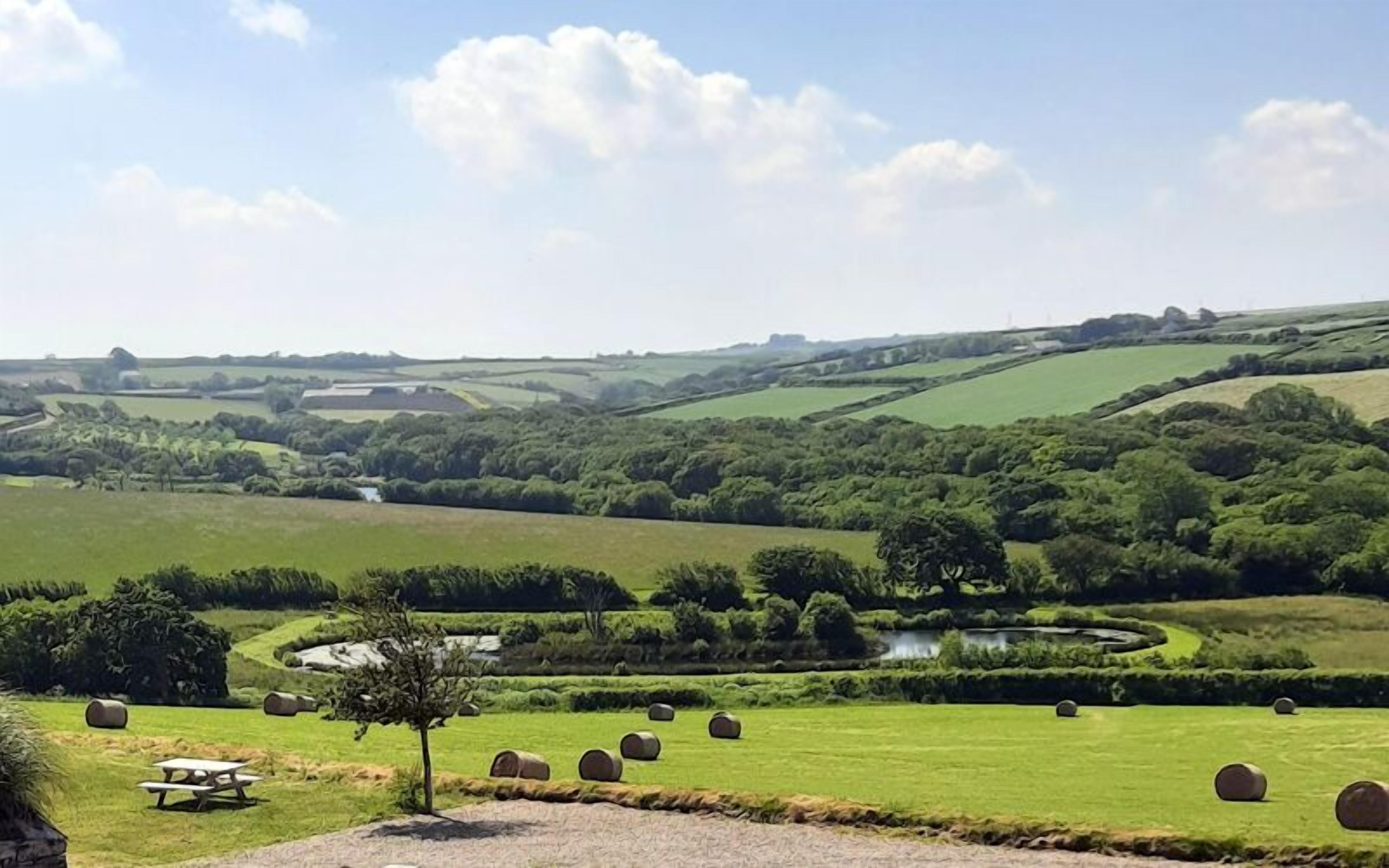 Paisaje rural con colinas onduladas, un pequeño estanque y pacas de heno dispersas por los campos.