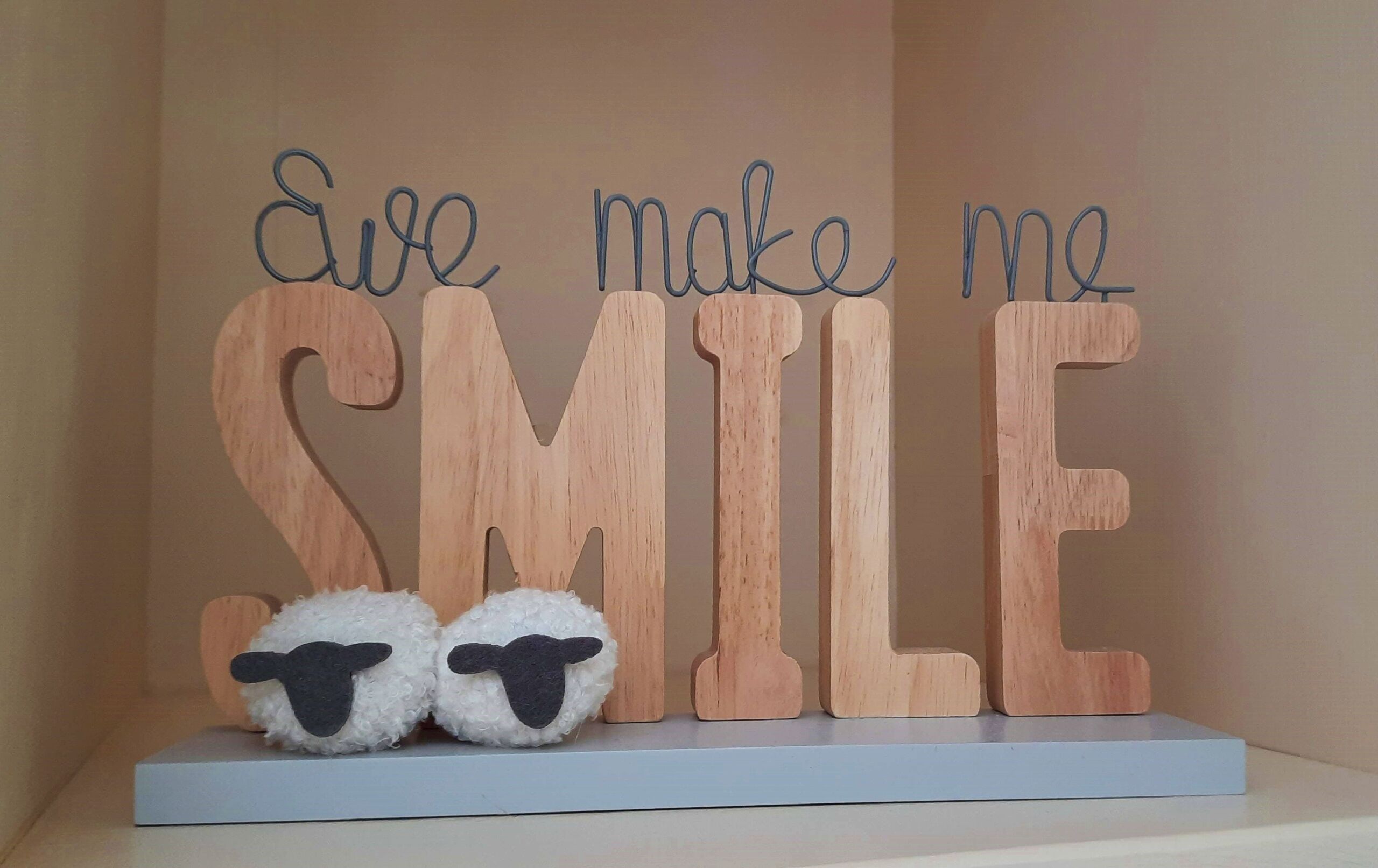 A wooden sign with the phrase "Eve make me SMILE" and two fluffy sheep figures on a shelf.
