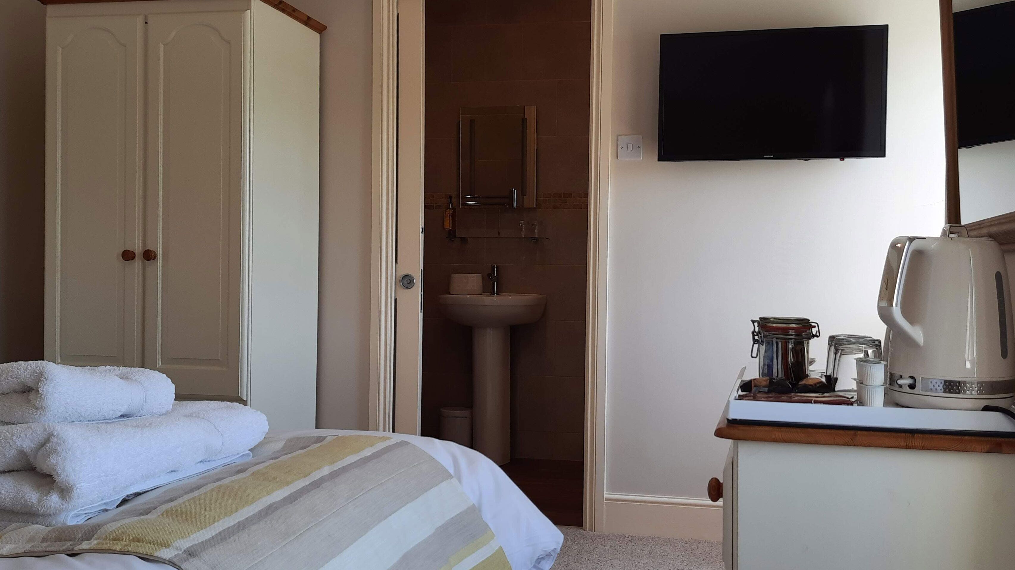 Une chambre avec un lit, une table de chevet avec une bouilloire, une télévision et une salle de bain avec un lavabo et un miroir.