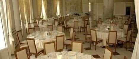 A banquet hall with round tables set for dining, chairs arranged around each table, and a large window providing natural light.