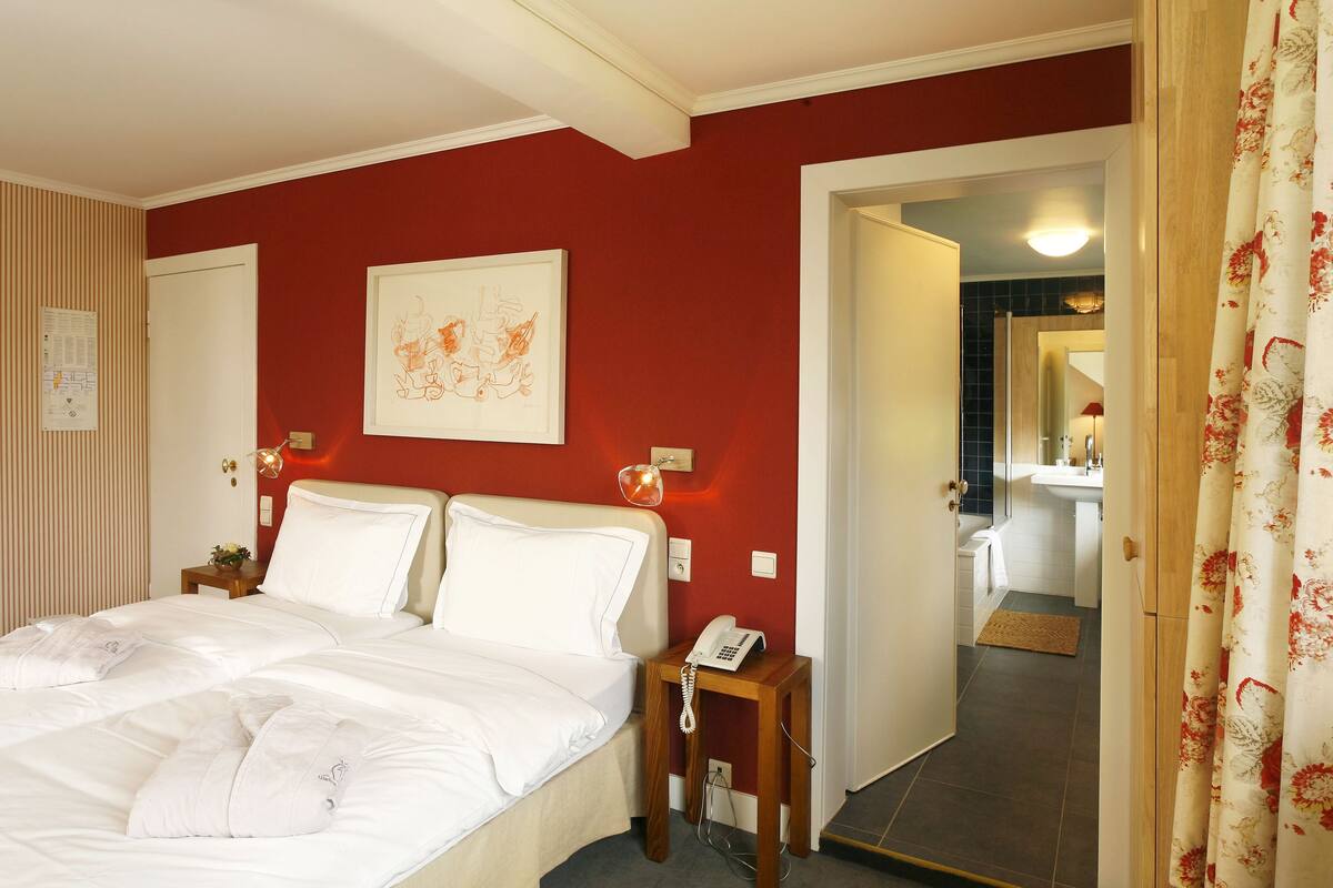 Een hotelkamer met een bed, een nachtkastje met een telefoon, een kunstwerk aan de muur en een badkamer zichtbaar door een open deur.
