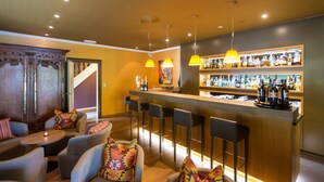 Een bar met een houten toonbank, barstoelen en diverse flessen. Er zijn zachte zitplekken met kleurrijke kussens.
