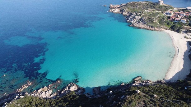 Vue aérienne d’une zone côtière avec une mer bleue et limpide, une plage de sable, un terrain rocheux et une structure fortifiée.