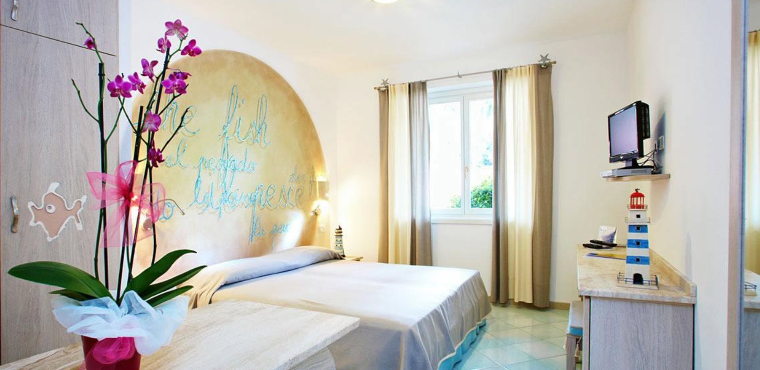 Habitación de hotel con cama, escritorio con computadora, ventana con cortinas y un muro decorativo con texto.