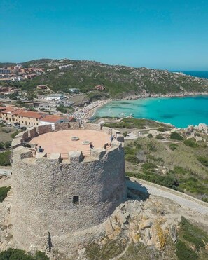 Une tour de pierre surmontée d’une structure circulaire, entourée d’un paysage côtier comprenant une plage et des eaux d’un bleu limpide.