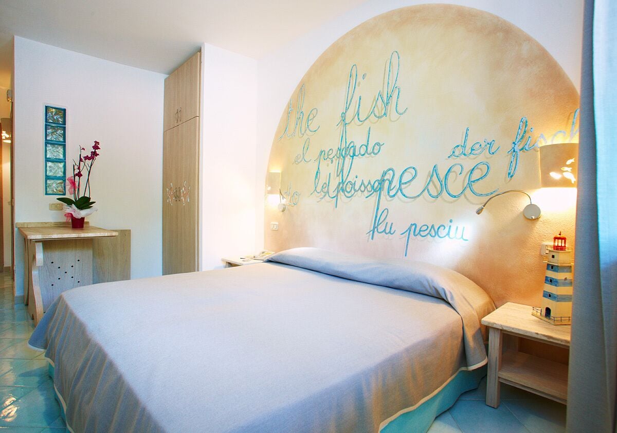 A bedroom with a bed, a nightstand, a wall clock, a small table with a vase of flowers, and a wall with text in multiple languages.