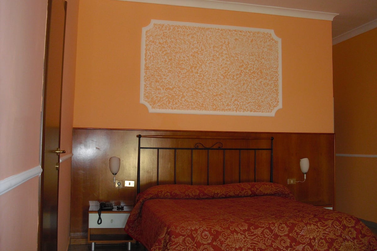 Habitación de hotel con cabecera de madera, cama con colcha estampada de color rojo, mesita de noche con lámpara y teléfono instalado en la pared.