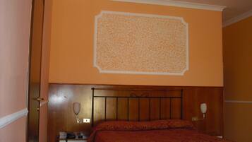 Ein Hotelzimmer mit einem hölzernen Kopfteil, einem Bett mit rot gemustertem Bettbezug, einem Nachttisch mit Lampe und einem an der Wand befestigten Telefon.