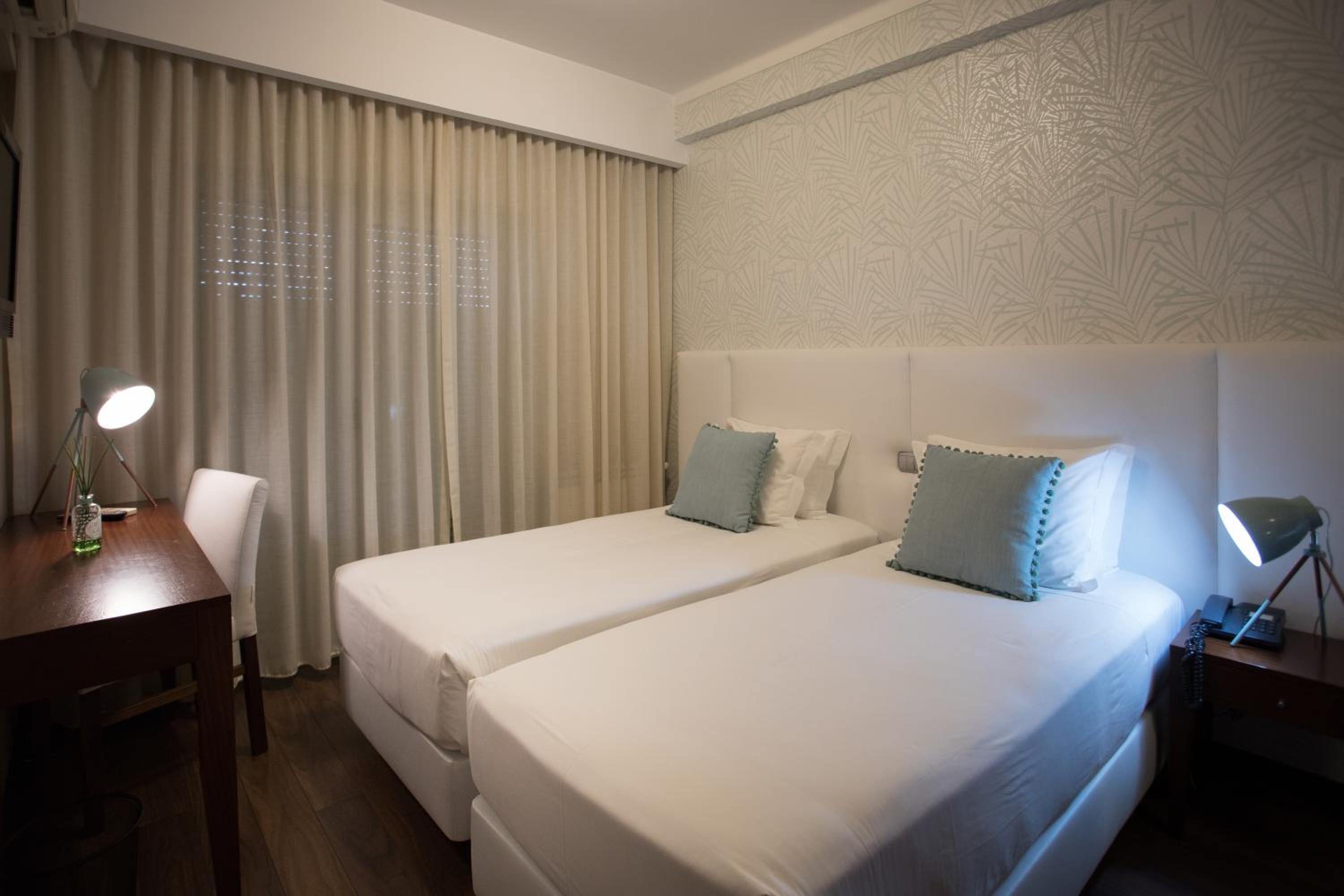 A hotel room with two single beds, a desk with a lamp, a chair, and a nightstand with a lamp.