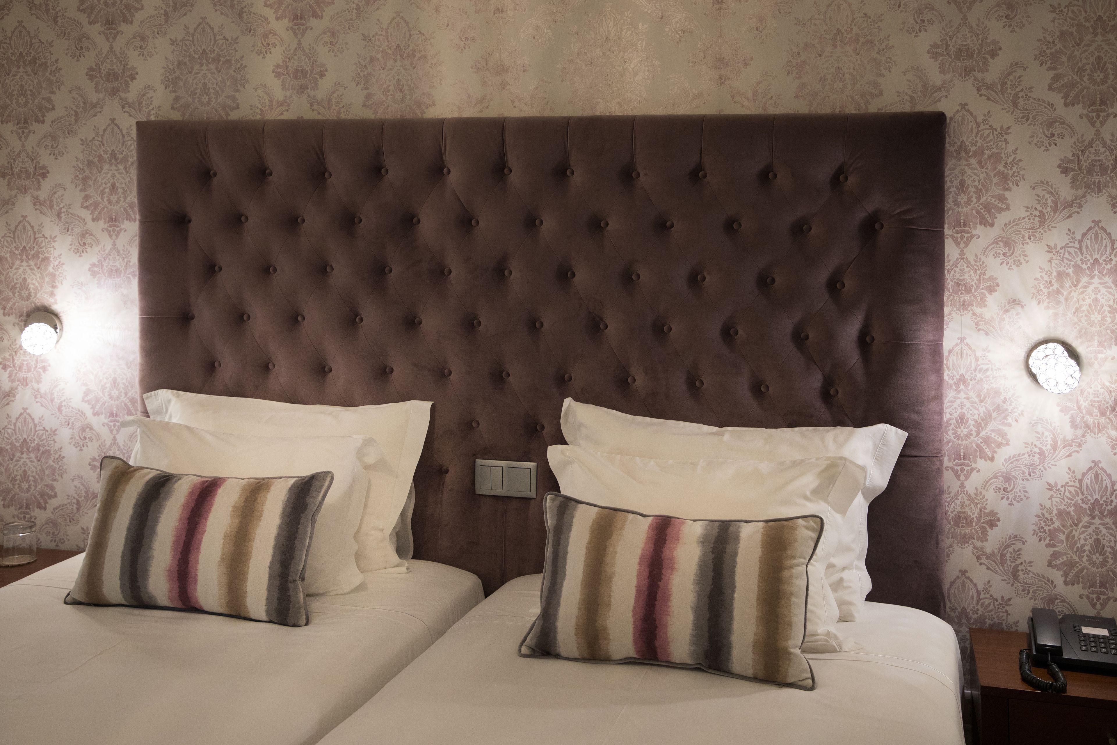Quarto de hotel com cabeceira estofada, duas camas com roupa de cama branca, almofadas listradas e telefone ao lado da cama.