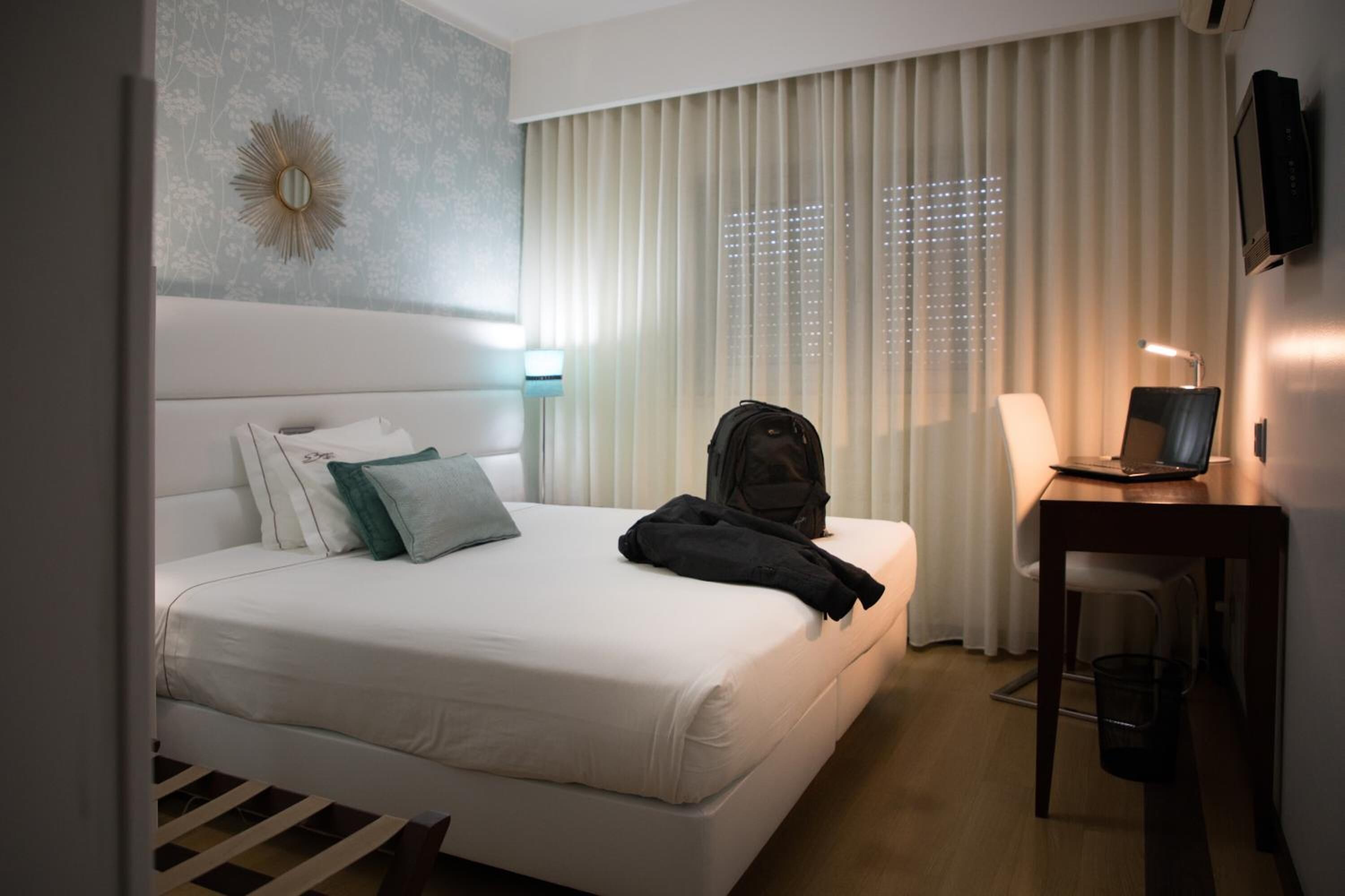 Een hotelkamer met een bed, een bureau met een laptop, een stoel, een televisie en een raam met jaloezieën.