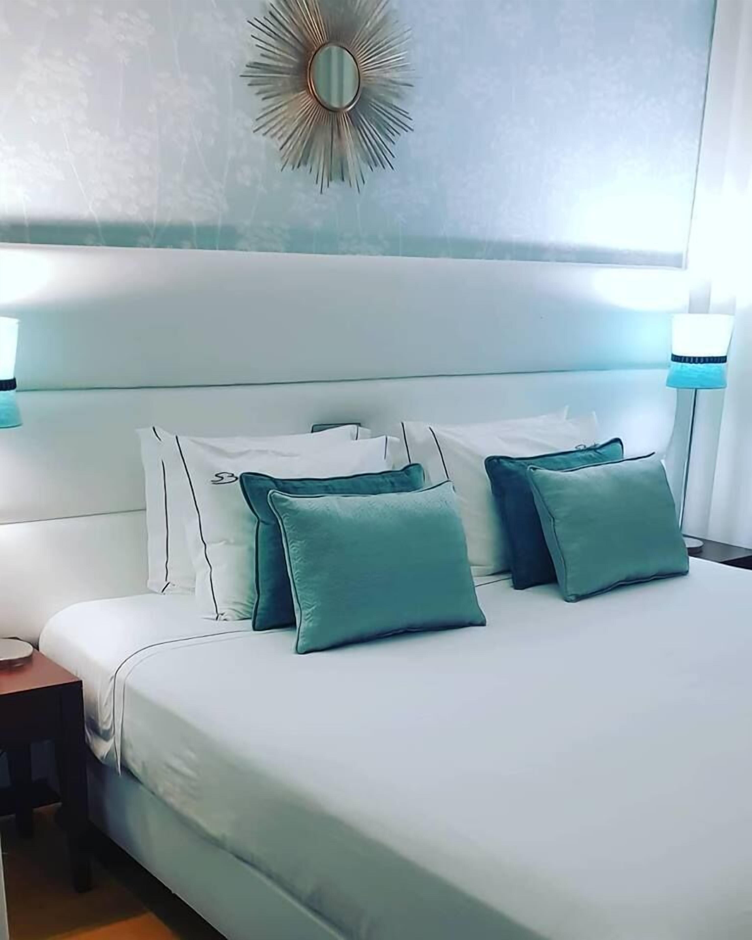 A neatly made bed with white and teal bedding, a round mirror on the wall, and a nightstand with a lamp.