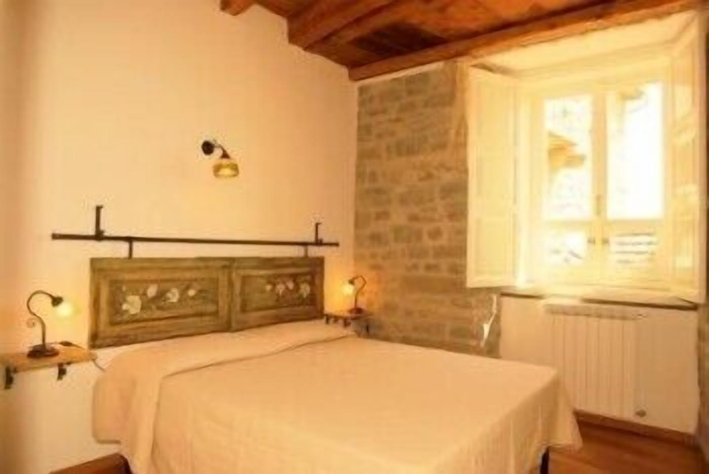 A bedroom with a wooden headboard, a bed with a white bedspread, two bedside lamps, a stone wall, and a window with a view of the outside.