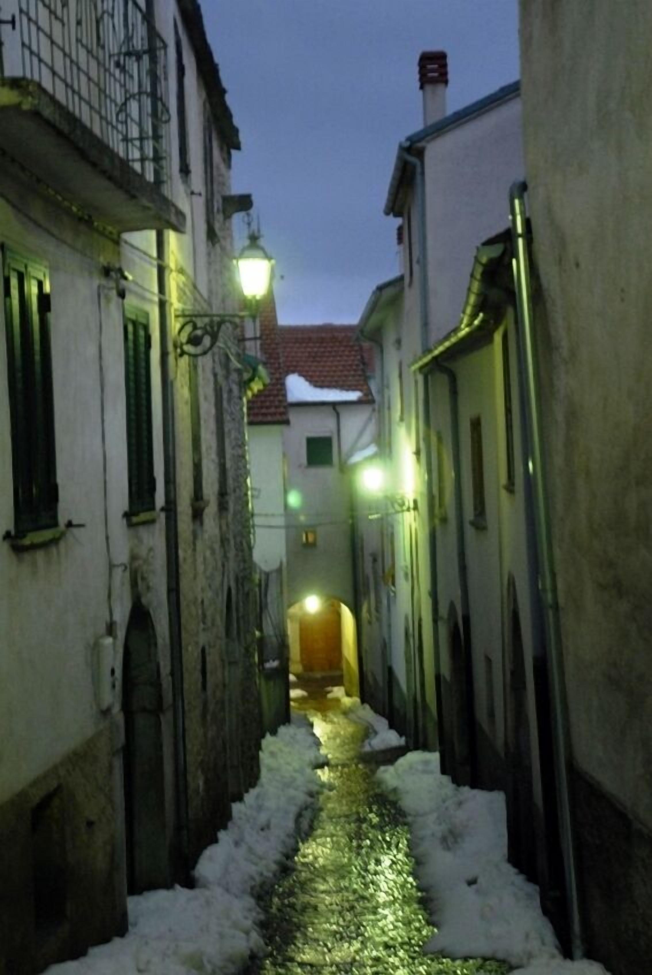 A narrow alleyway with snow on the ground, flanked by buildings with visible windows and doors.