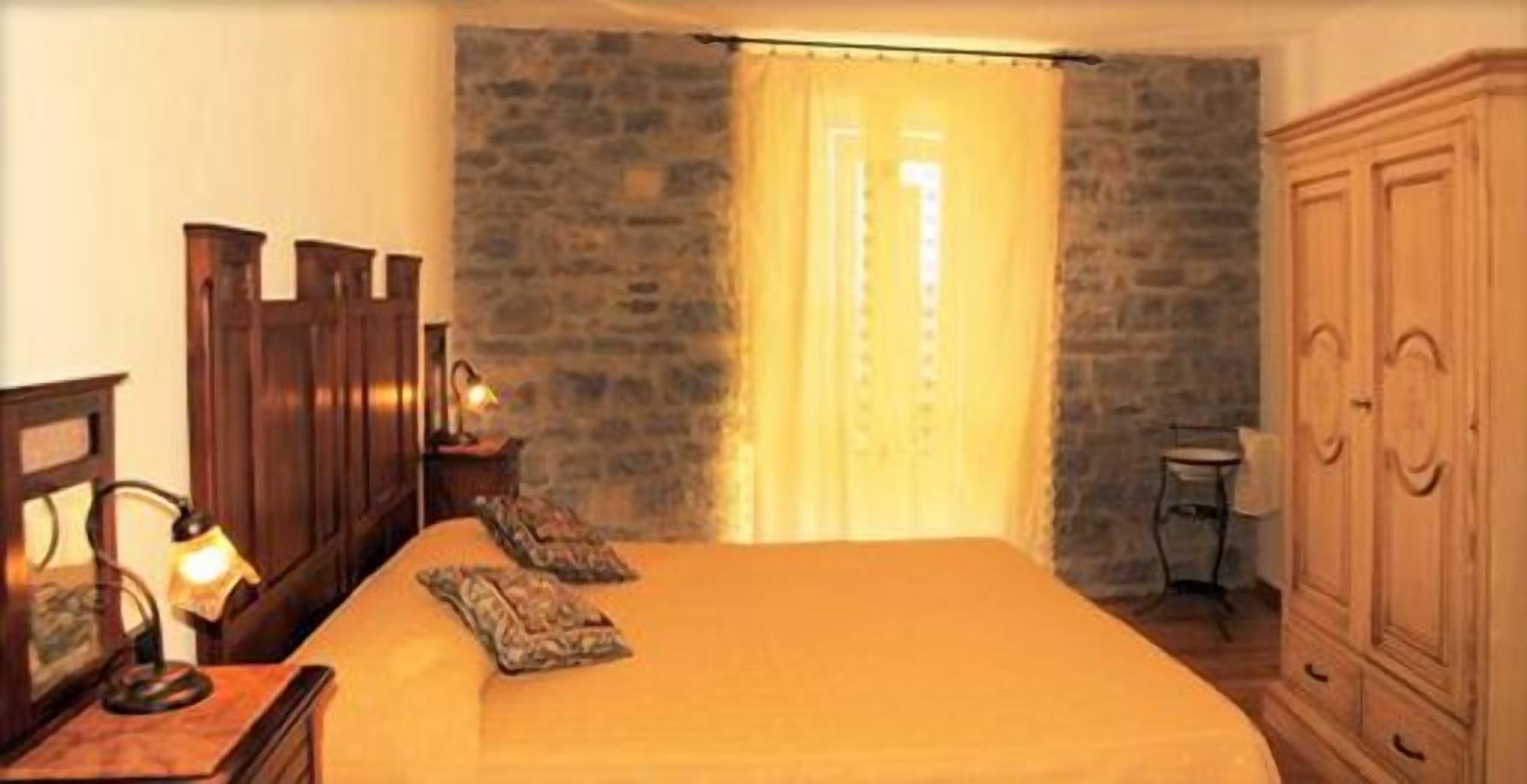 A bedroom with a bed, wooden wardrobe, and a stone wall.
