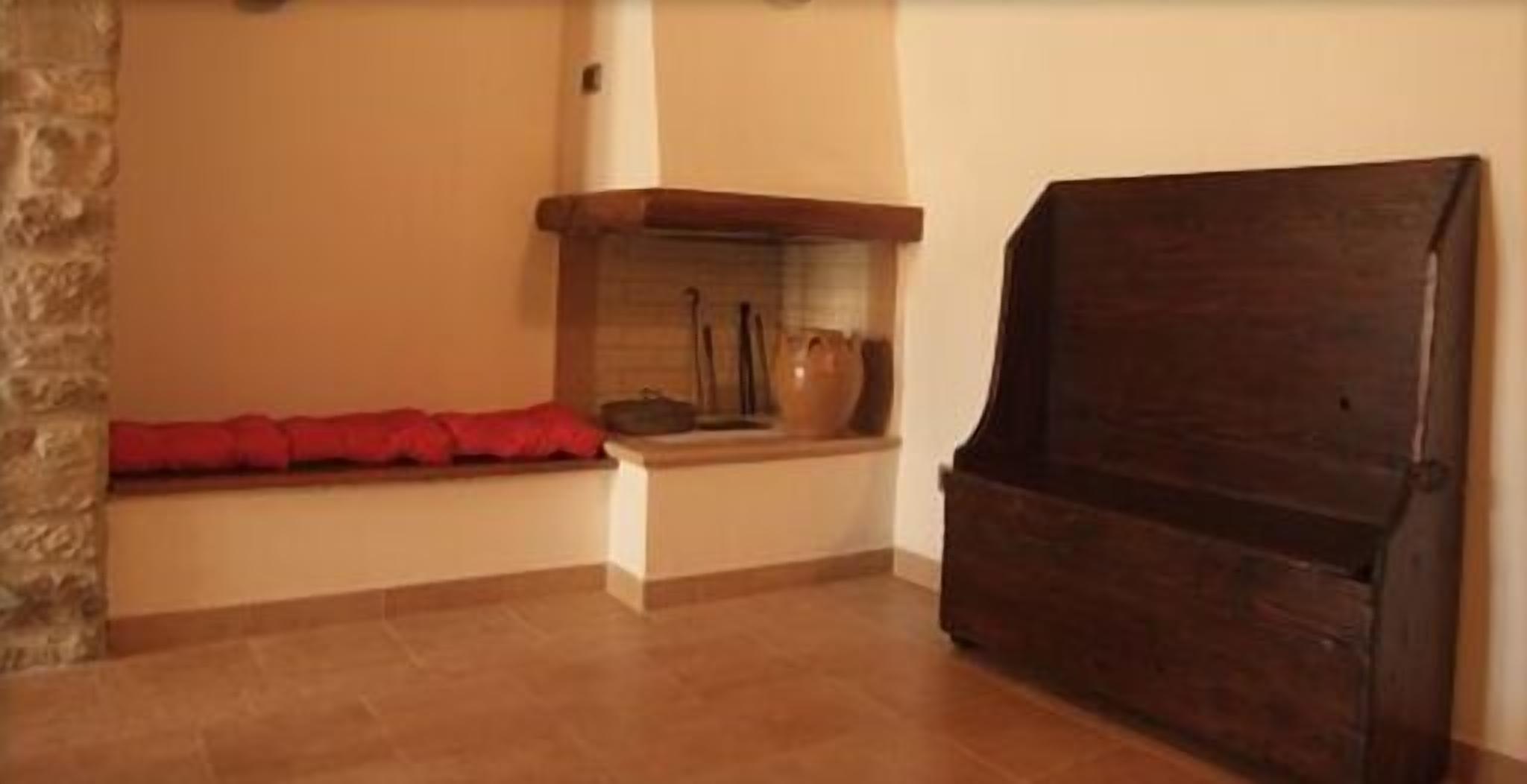 Una habitación con pared de piedra, un banco de madera con cojines rojos, una chimenea y un armario de madera oscura.