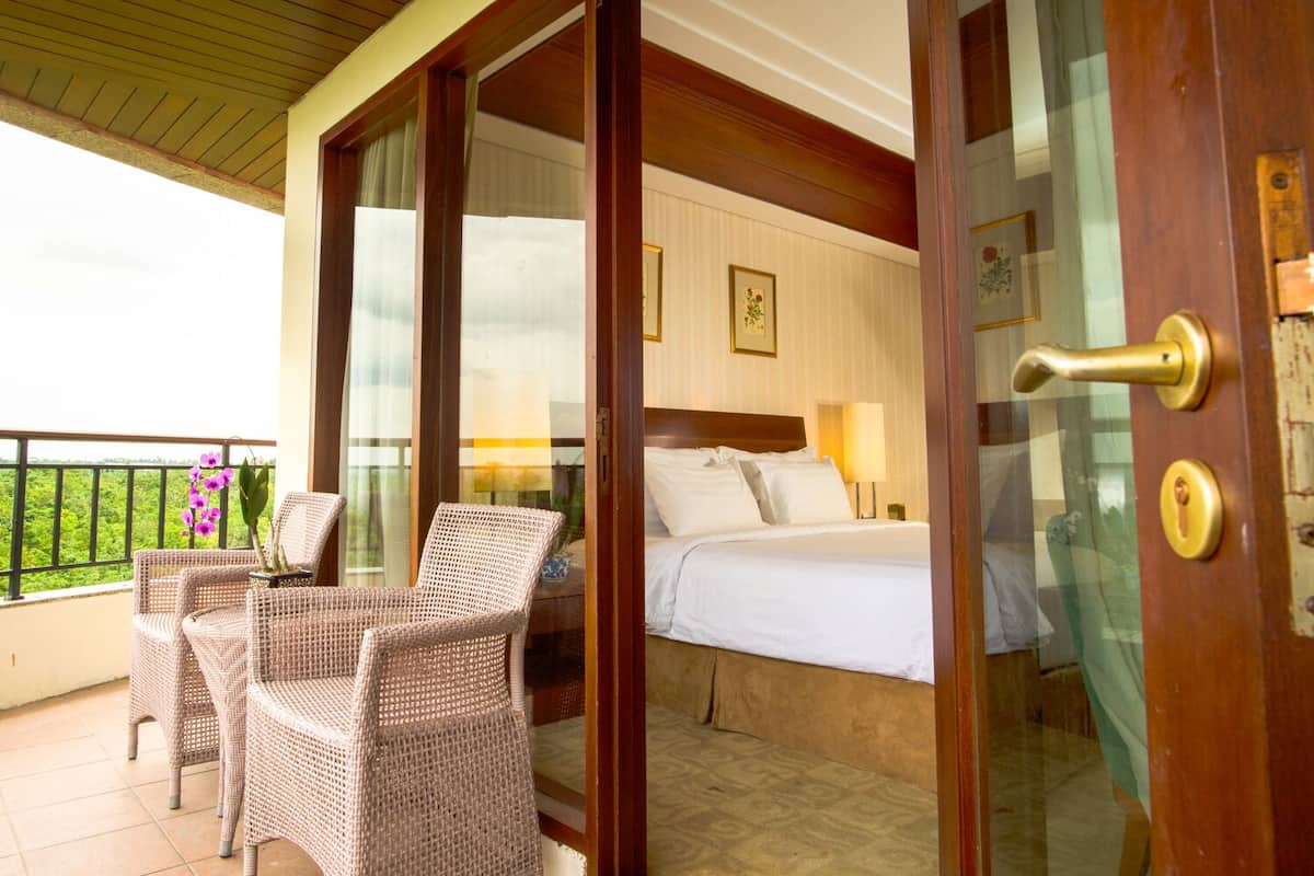 A room with a bed, a balcony with wicker chairs and a potted orchid, and a view of greenery.