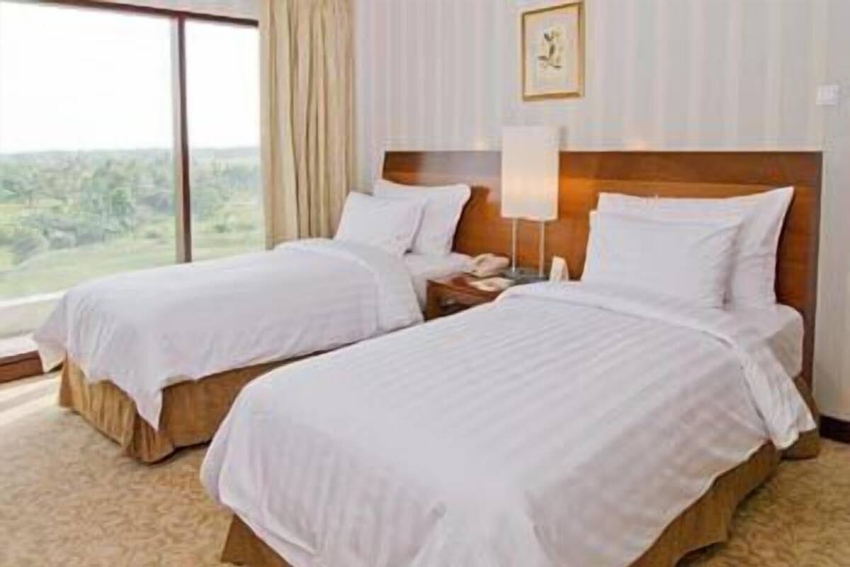 A hotel room with two beds, a nightstand, a lamp, and a window with a view of greenery.