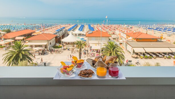 A balcony with breakfast items and a view of a beach with umbrellas and beach chairs. - Giulia Hotel (Camaiore)