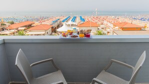 A balcony with wicker chairs, a table with breakfast items, and a view of a beach with umbrellas and beach chairs. - Giulia Hotel (Camaiore)