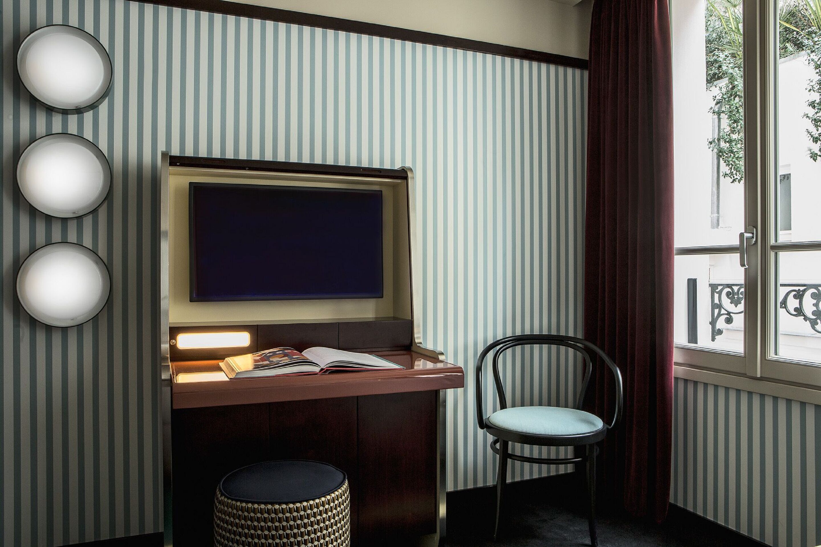 A room with a television, a desk with a book, a chair, and a window with a view of the outside.