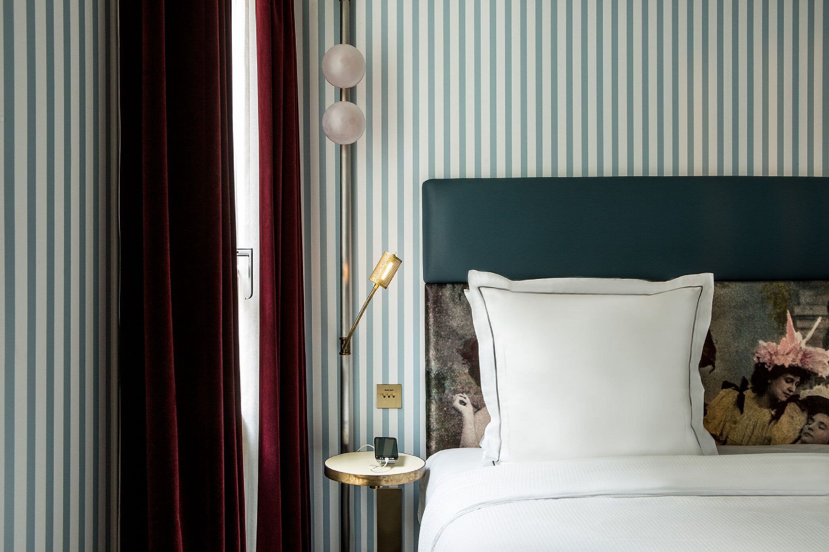 Une chambre d’hôtel avec un papier peint à rayures, une tête de lit sombre, une table de chevet avec une lampe et un tableau au mur.