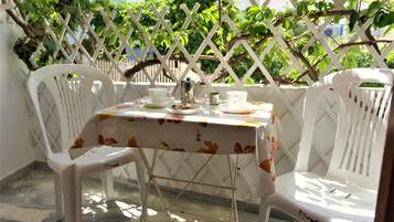 A small outdoor table with a floral tablecloth, two white plastic chairs, and a white lattice fence in the background.