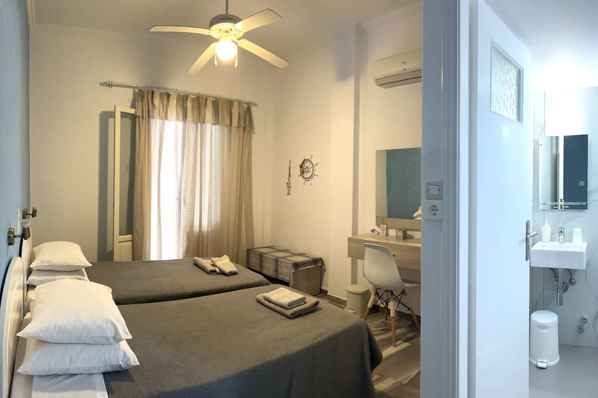 Habitación de hotel con dos camas, un escritorio, una silla, un espejo y un baño con lavamanos e inodoro.