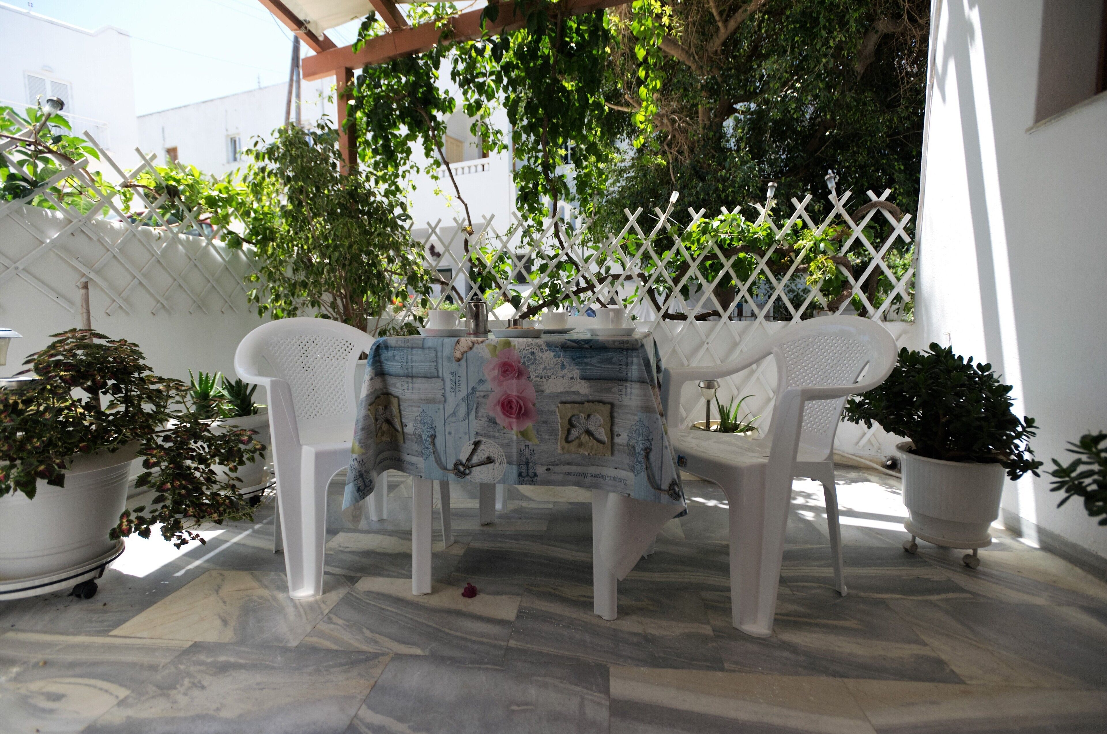 A patio with a table covered by a tablecloth, surrounded by potted plants and white chairs.