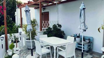 An outdoor patio with white tables and chairs, umbrellas, potted plants, and a pergola.