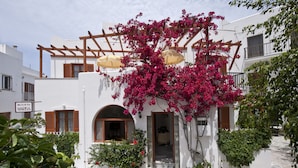 Un edificio blanco con una pérgola de madera, buganvillas rosadas y un letrero que dice "ROOMS MARISA".