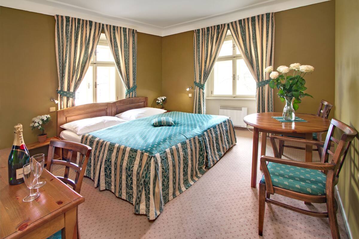 Ein Hotelzimmer mit einem Bett, einem Holztisch mit einer Champagnerflasche, zwei Stühlen, einer Blumenvase und Vorhängen.