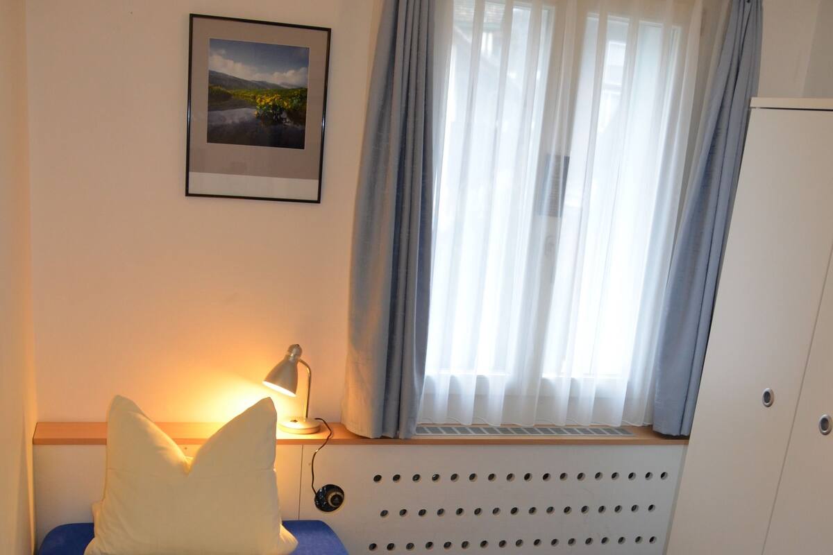 A single bed with a blue mattress and white bedding, a bedside lamp, a radiator, a window with curtains, and a wardrobe.